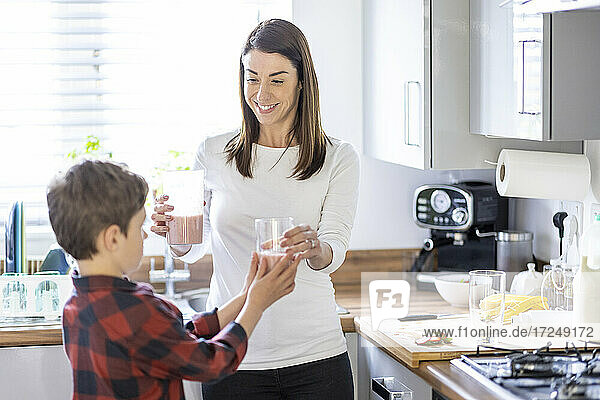 Smiling mother giving glass of milkshake to son in kitchen at home