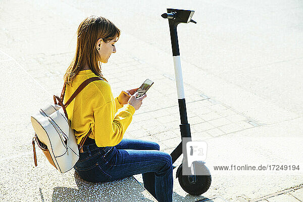 Junge Frau  die ein Mobiltelefon benutzt  während sie auf einer Bank mit einem Elektroroller sitzt