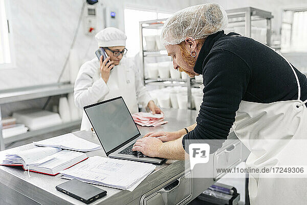 Besitzer benutzt Laptop  während ein Kollege im Hintergrund in der Fabrik arbeitet