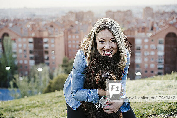 Lächelnde Frau mit blondem Haar umarmt spanischen Wasserhund auf Gras bei Sonnenuntergang