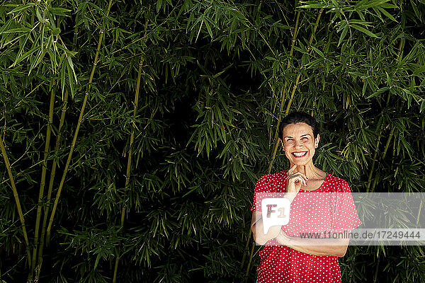 Fröhliche Frau mit Hand am Kinn vor Bambuspflanzen stehend