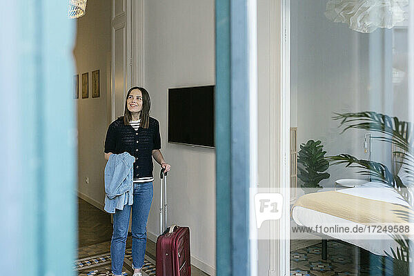 Smiling young woman with suitcase looking away while standing in apartment