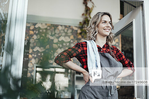 Glückliche Frau mit Hand an der Hüfte  die wegschaut  während sie am Eingang eines Cafés steht