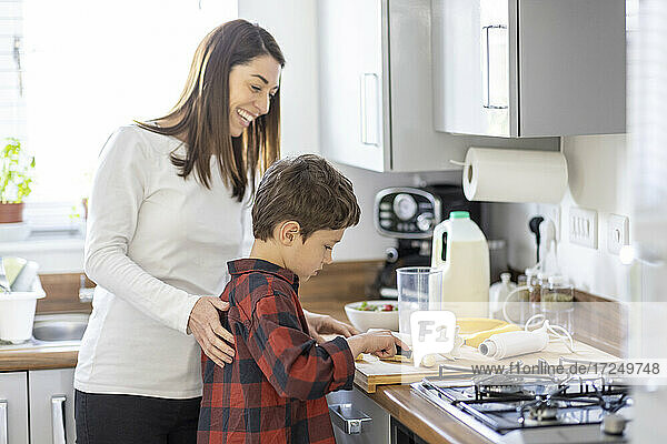 Lächelnde Mutter hilft ihrem Sohn beim Schneiden von Bananen in der Küche zu Hause