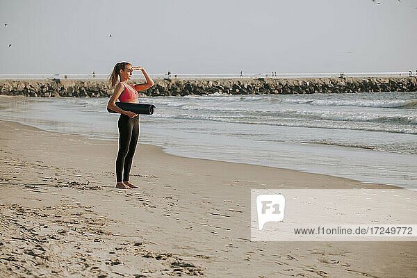 Junge Frau  die ihre Augen abschirmt  während sie eine Übungsmatte am Strand hält