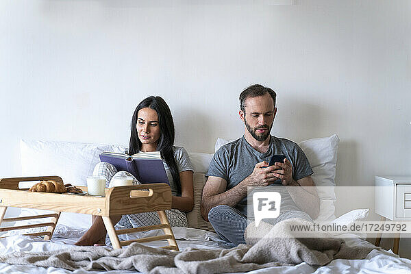 Frau,  die ein Buch liest und mit einem Mann zusammensitzt,  der ein Smartphone benutzt,  während sie zu Hause auf dem Bett frühstückt
