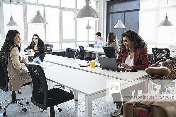 Lächelnde Unternehmerinnen  die am Schreibtisch in einem Coworking-Büro diskutieren