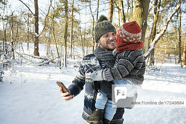 Father carrying son while holding smart phone in forest during winter