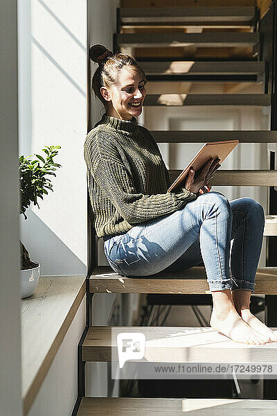 Lächelnde Frau  die ein Tablet benutzt  während sie auf einer Treppe zu Hause sitzt