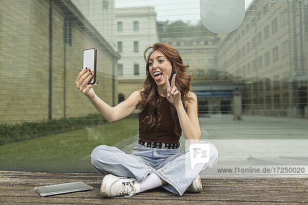 Frau gestikuliert bei der Aufnahme eines Selfies mit dem Handy vor einer Glaswand