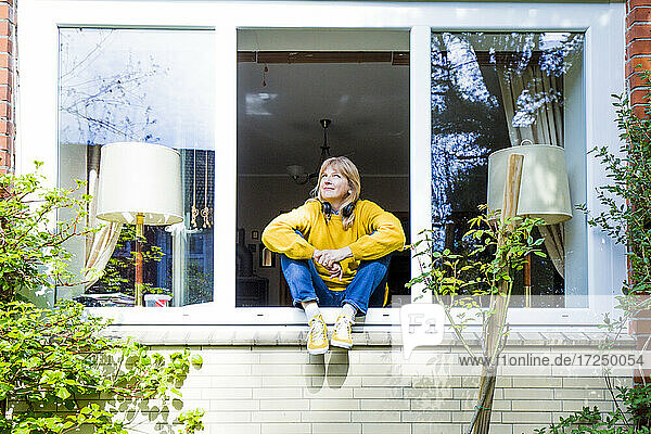 Reife Frau blickt nach oben  während sie zu Hause auf der Fensterbank sitzt