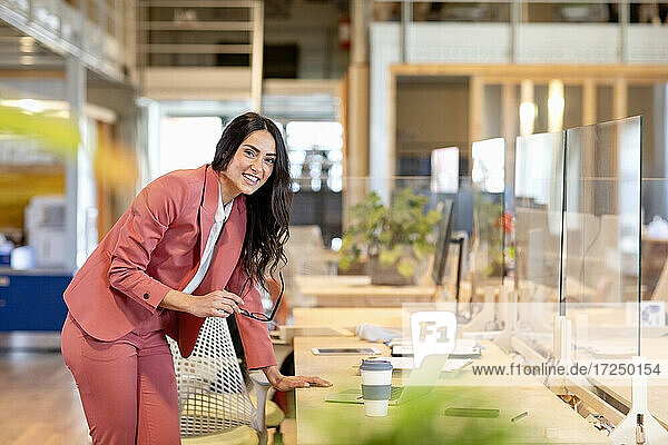 Lächelnde weibliche Fachkraft lehnt sich an einen Schreibtisch in einem Coworking-Büro