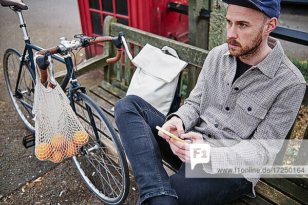 Thoughtful man with mobile phone sitting on bench by bicycle