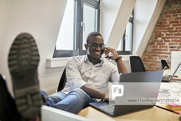 Männlicher Unternehmer  der mit seinem Smartphone spricht  während er einen Laptop im Büro benutzt