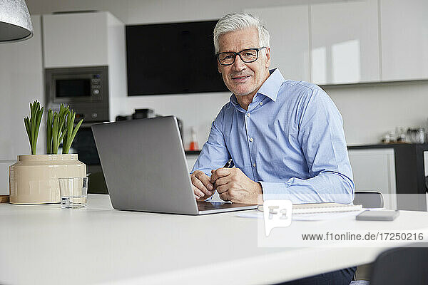 Lächelnder Geschäftsmann mit Laptop sitzt am Schreibtisch im Büro