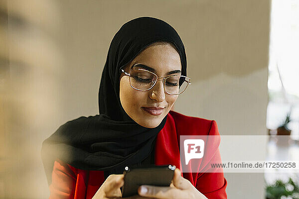 Frau mit Hidschab lächelt  während sie in einem Café ein Mobiltelefon benutzt