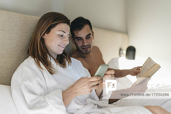 Beautiful woman using phone while sitting by man with book on bed
