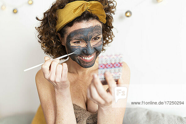 Cheerful woman applying face mask in bedroom at home
