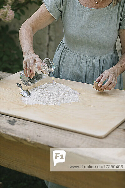 Reife Frau  die Wasser auf Mehl gießt  während sie Teig im Vorgarten oder Hinterhof zubereitet