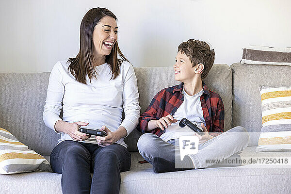 Fröhliche Frau sieht ihren Sohn an  während sie auf dem Sofa im Wohnzimmer ein Videospiel spielt