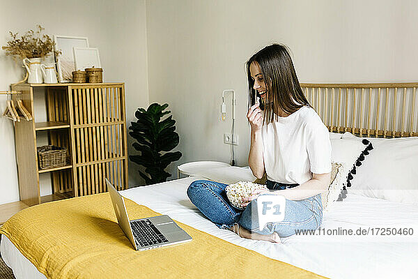 Frau isst Popcorn  während sie zu Hause auf dem Bett ihren Laptop betrachtet