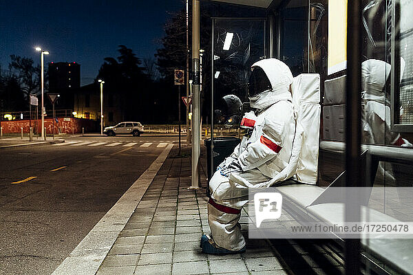 Young female astronaut in space suit waiting at bus stop during night