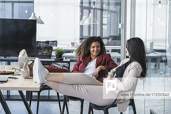 Lächelnde Geschäftsfrauen  die mit hochgelegten Füßen auf einem Stuhl in einem Coworking-Büro sitzen und diskutieren