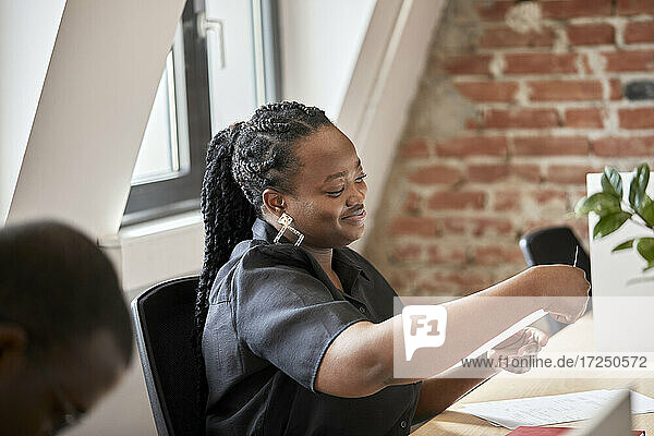 Lächelnde Geschäftsfrau mit geflochtenem Haar  die ein Dokument in einem Coworking-Büro betrachtet