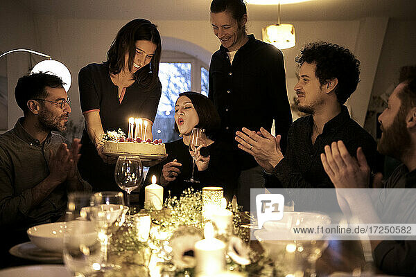 Frau bläst Kerzen auf einem Geburtstagskuchen aus  während männliche und weibliche Freunde bei einer Feier zu Hause klatschen