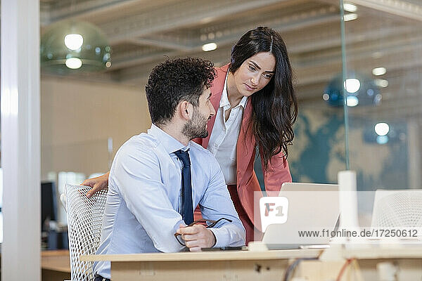 Female professional looking at laptop by male colleague at desk in coworking office