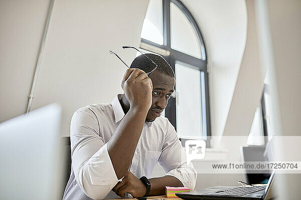 Besorgter männlicher Berufstätiger  der eine Brille hält und auf einen Laptop im Büro schaut