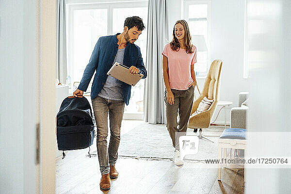 Smiling young woman looking at businessman carrying baby carrier in living room