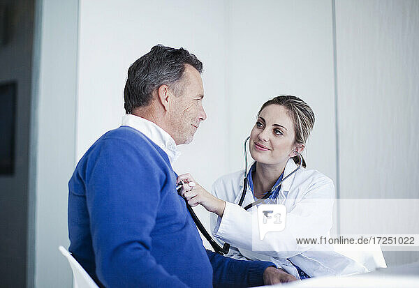 Smiling female doctor assessing male patient with stethoscope in hospital