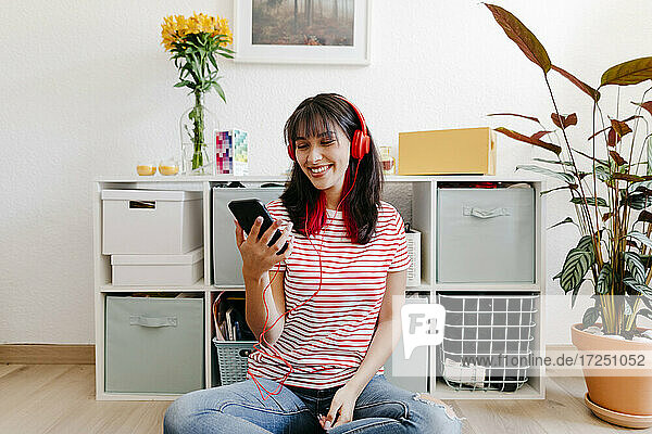 Lächelnde Frau mit Kopfhörern  die ein Smartphone benutzt  während sie zu Hause sitzt