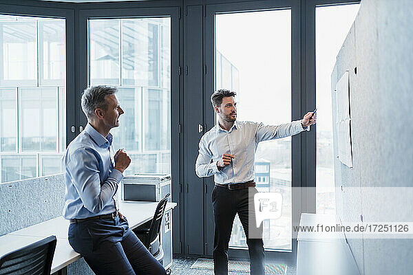 Businessman explaining paper over bulletin board with male colleague in office
