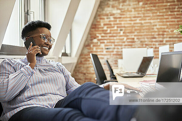 Weibliche Fachkraft mit Brille  die mit einem Smartphone spricht  während sie einen Laptop im Büro benutzt