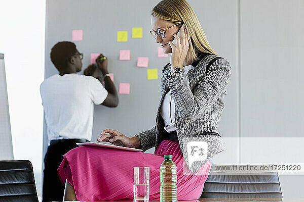 Geschäftsfrau  die mit einem Smartphone spricht  während ein männlicher Kollege im Hintergrund im Büro arbeitet
