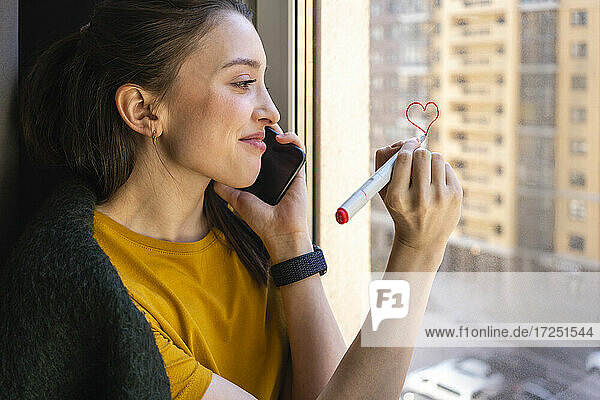 Young woman drawing heart while talking on mobile phone at home