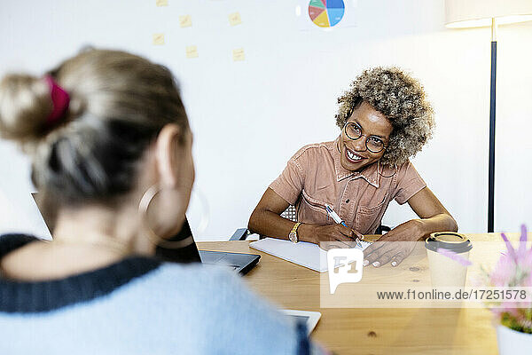 Businesswoman discussing with female colleague while writing in book at home office