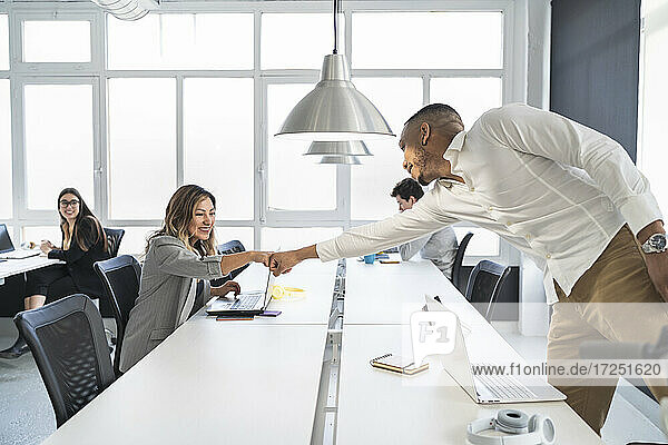 Lächelnde männliche und weibliche Unternehmer  die sich am Schreibtisch in einem Coworking-Büro die Fäuste stoßen