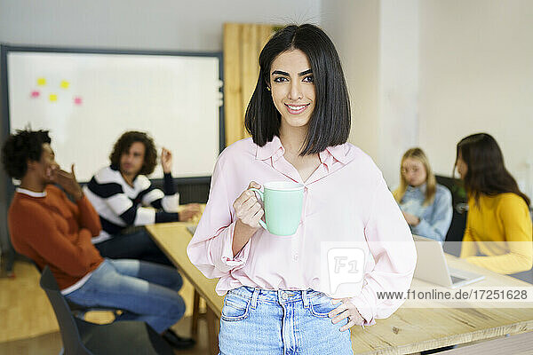 Weibliche Fachkraft mit Hand auf der Hüfte  die einen Kaffeebecher hält  während sie sich an einen Schreibtisch lehnt