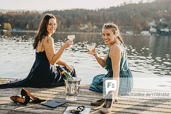 Lächelnde Veranstaltungsplanerinnen sitzen mit Champagner auf einem Steg über dem See