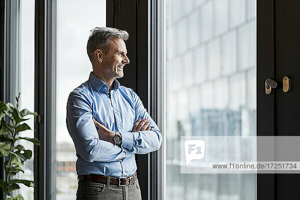 Smiling businessman with arms crossed day dreaming while standing at work place