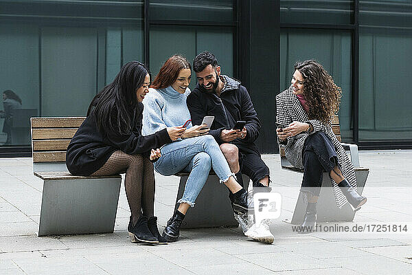 Junger Mann und Frauen benutzen ein Mobiltelefon  während sie auf einer Bank sitzen