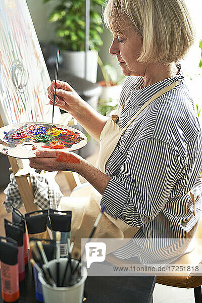 Blonde Frau mit Pinsel mischt Farben auf einer Palette vor einer Staffelei zu Hause