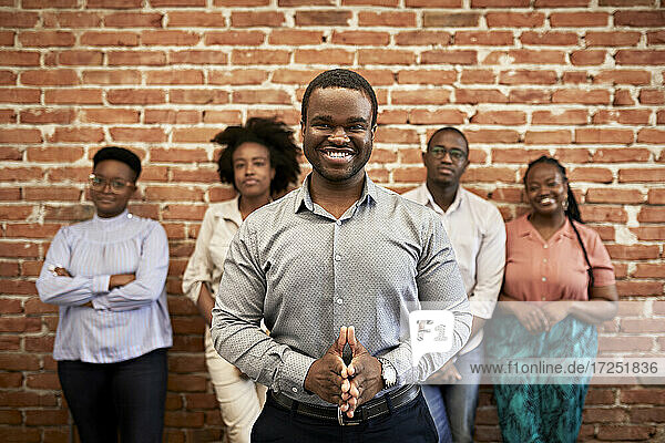 Lächelnder männlicher Berufstätiger mit verschränkten Händen  im Hintergrund stehen Kollegen im Büro