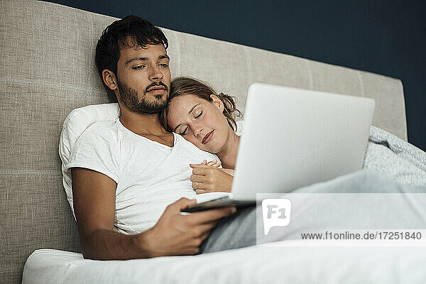 Junger Mann benutzt Laptop bei seiner Freundin  die zu Hause auf dem Bett schläft