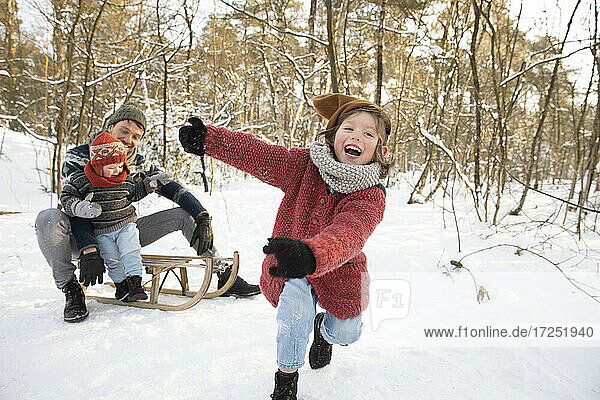 Verspielter Junge im Schnee  während der Vater mit dem jüngeren Sohn auf dem Schlitten im Hintergrund sitzt