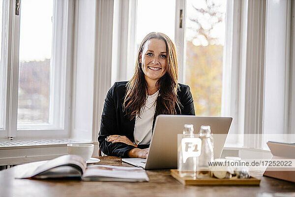 Lächelnde Veranstaltungsplanerin sitzt mit Laptop am Tisch im Büro