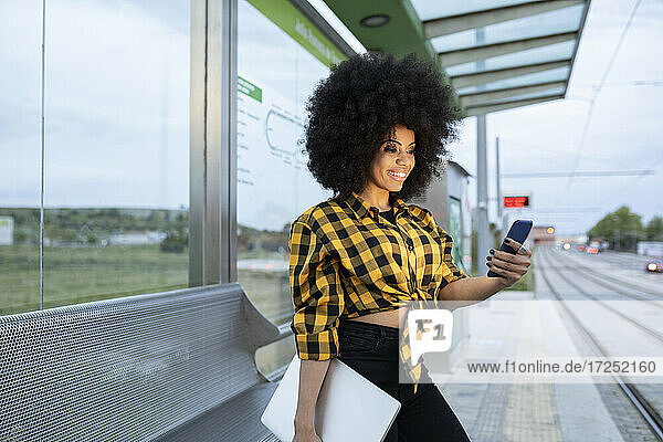 Lächelnde Frau  die einen Laptop hält und ein Smartphone am Bahnhof benutzt